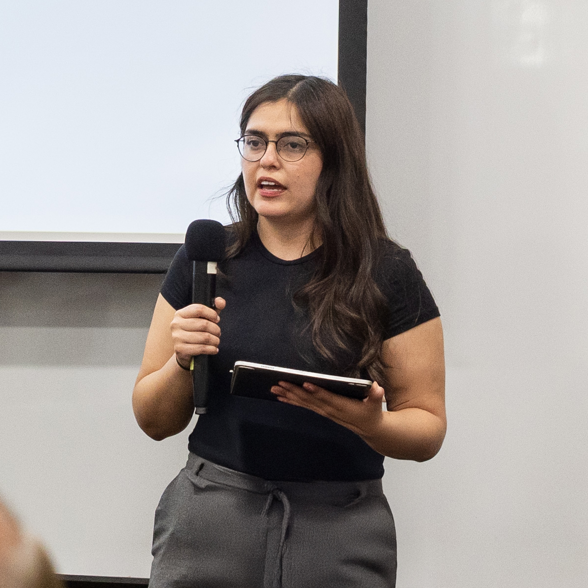 An MSBA student holding a microphone and iPad, in front of a projection screen, giving a presentation.